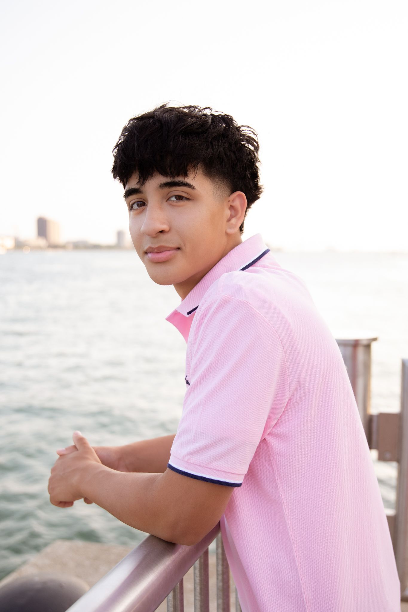 Young man in pink polo shirt leaning on railing by waterfront, smiling over shoulder with city skyline in background.