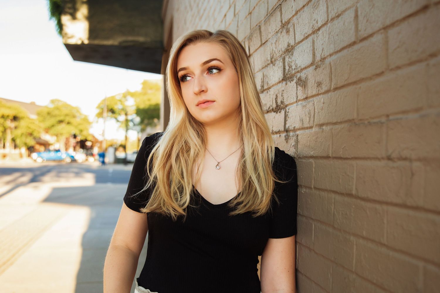 Young blonde woman in black t-shirt leaning against brick wall outdoors in golden hour sunlight.