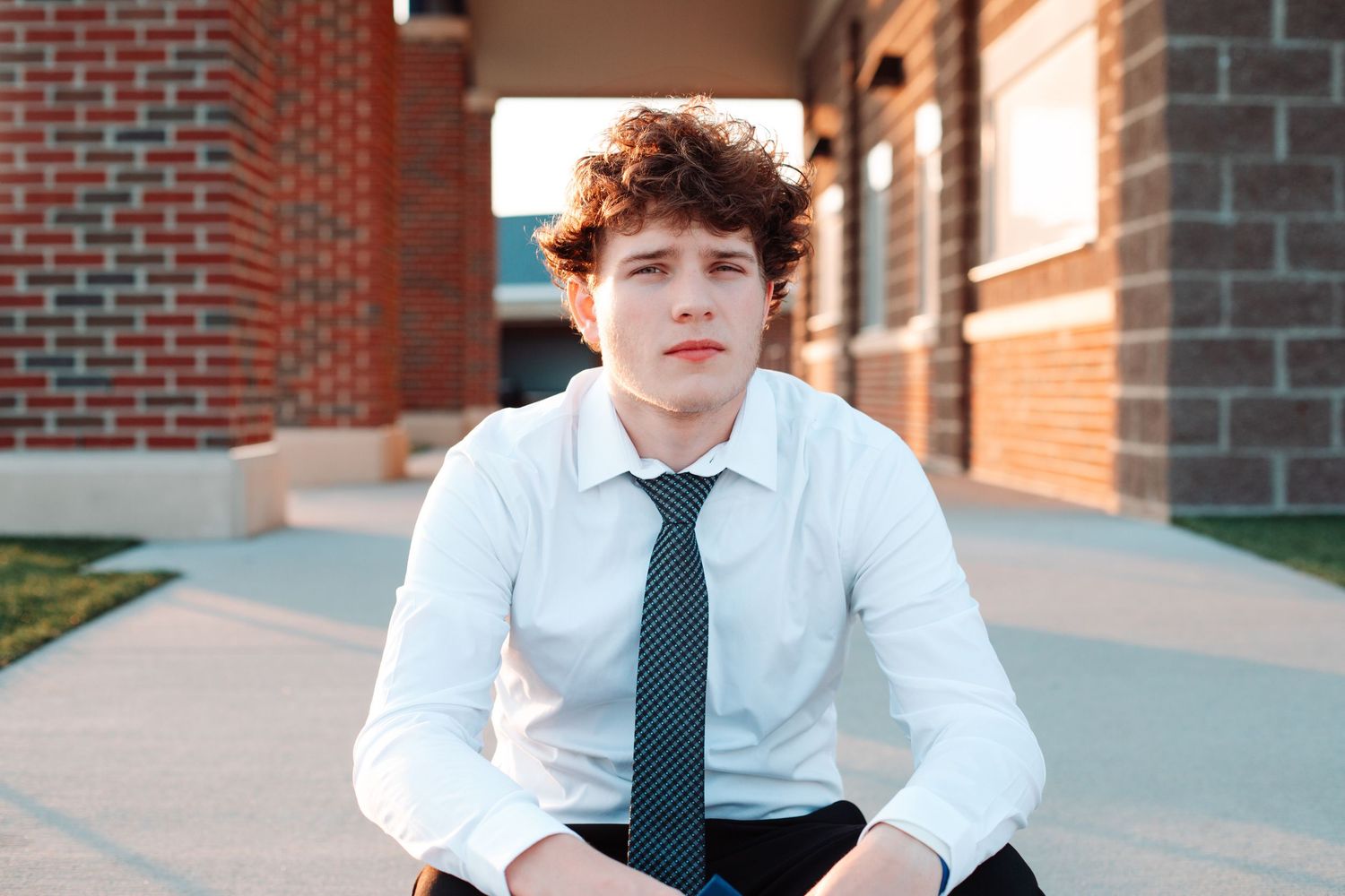 Young man in white dress shirt and tie sitting outdoors near brick building for senior portrait photo.