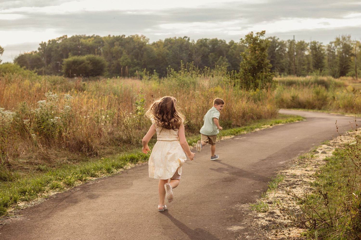 Two children running along a scenic path surrounded by tall grass and trees on a warm summer day.