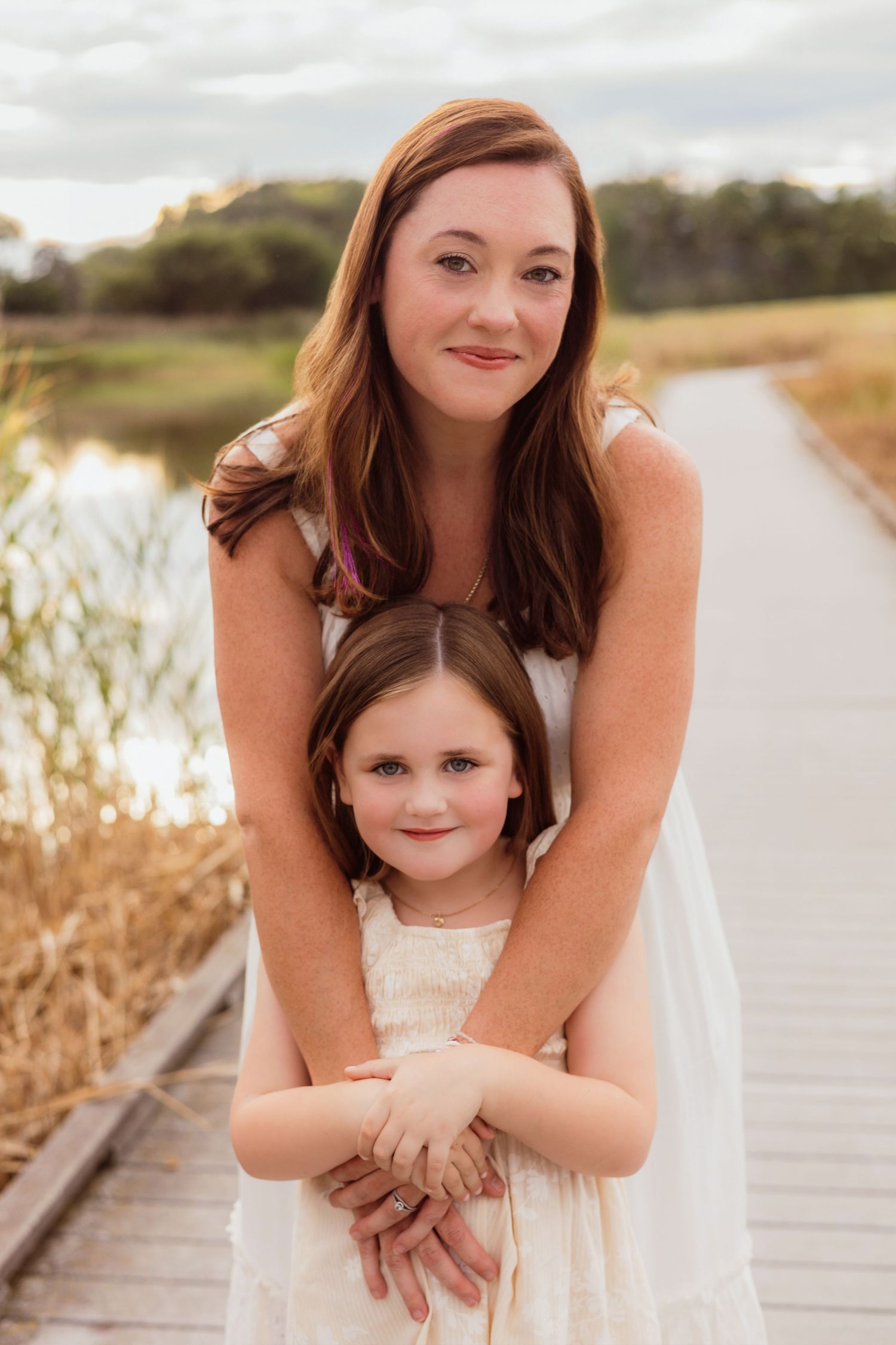 A woman in a white dress hugs a young girl on a wooden boardwalk near a lake at golden hour.