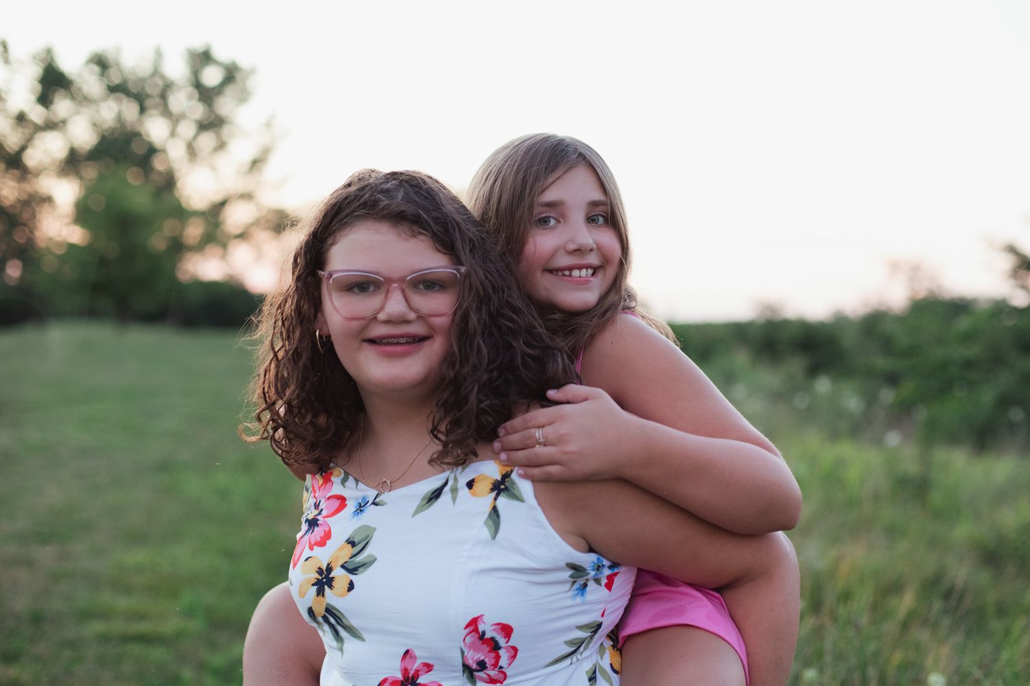 Two smiling girls pose outdoors at sunset, one giving the other a piggyback ride in a green field.