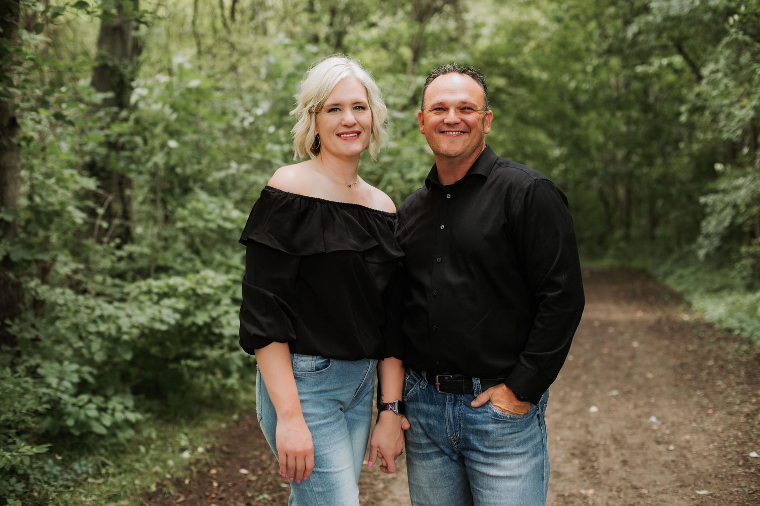 Smiling couple in black outfits posing on a scenic wooded path.