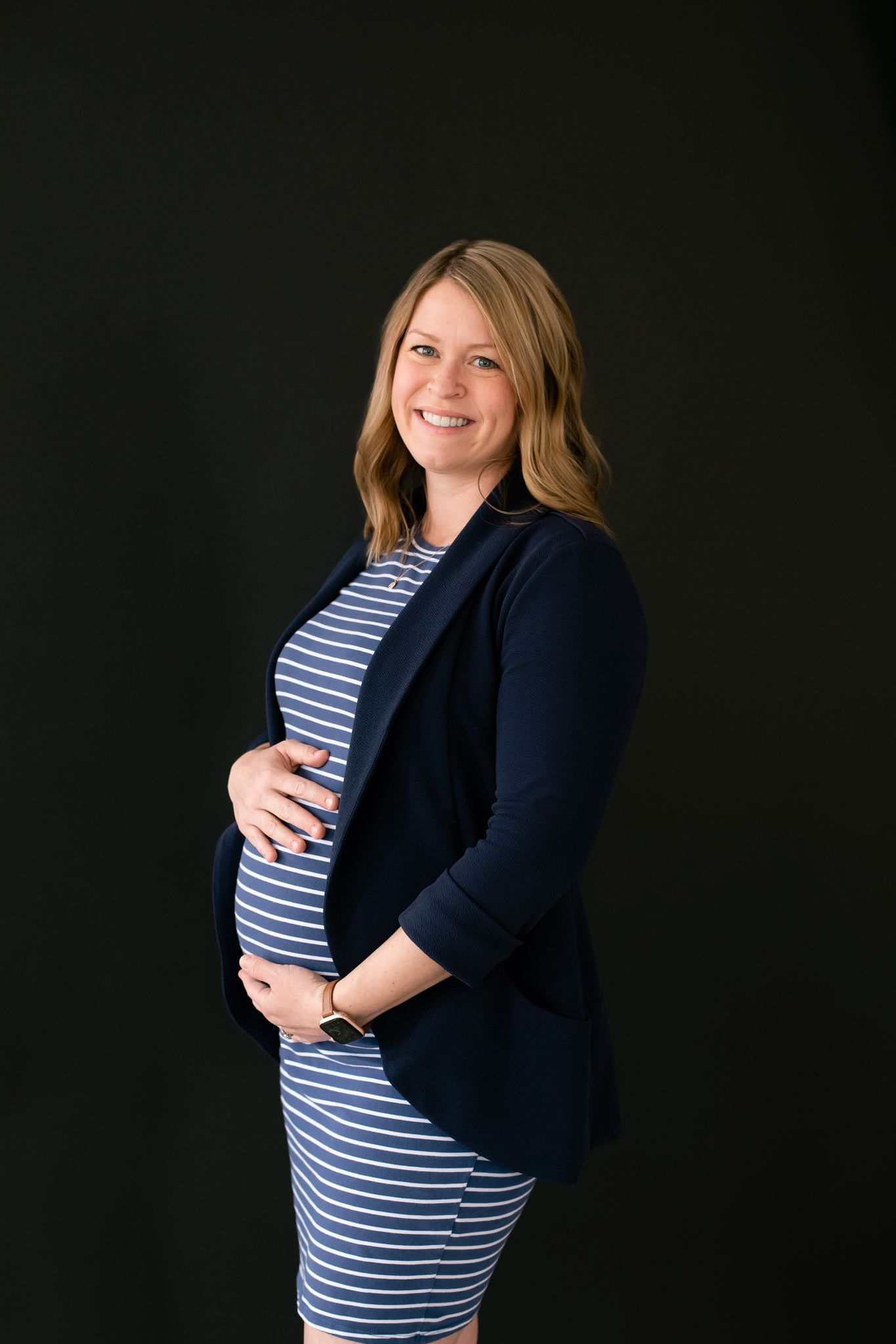 Smiling pregnant woman in striped dress and navy blazer cradles her baby bump against dark background.
