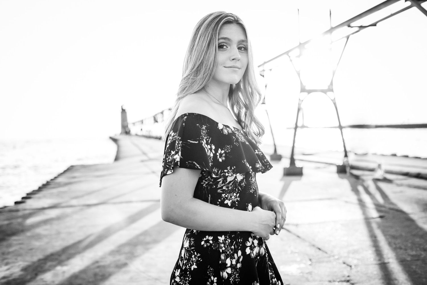 Black and white portrait of a blonde woman in a floral dress on a waterfront pier.