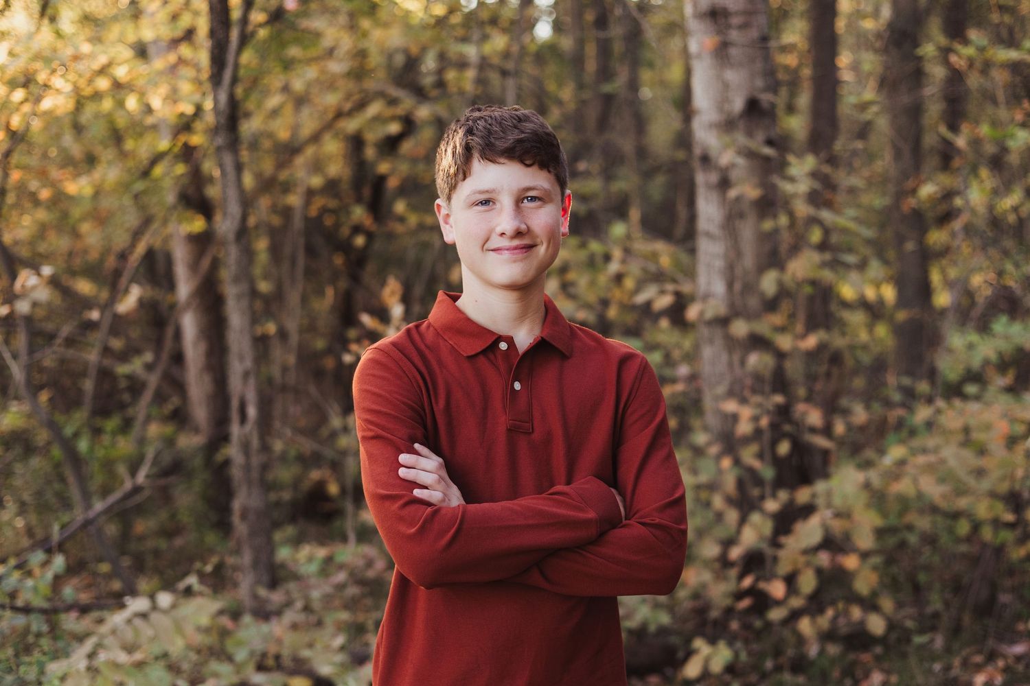 Smiling teenage boy in red polo shirt posing with arms crossed among colorful fall foliage.