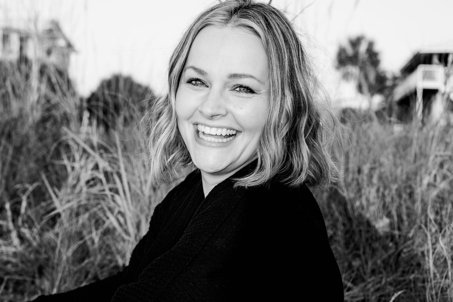 Smiling blonde woman in black outfit posing outdoors among tall grasses in a black and white portrait photo.