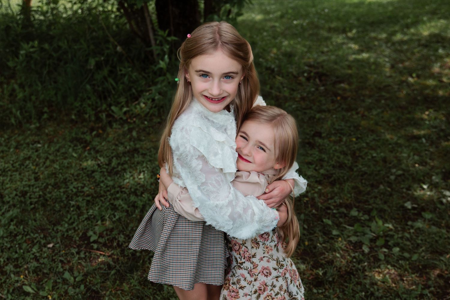 Two young girls with long hair hugging and smiling outdoors in a lush green garden setting.