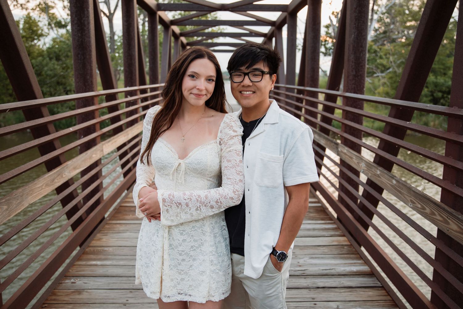 Engaged couple posing together on a wooden bridge wearing white outfits during outdoor engagement photos.