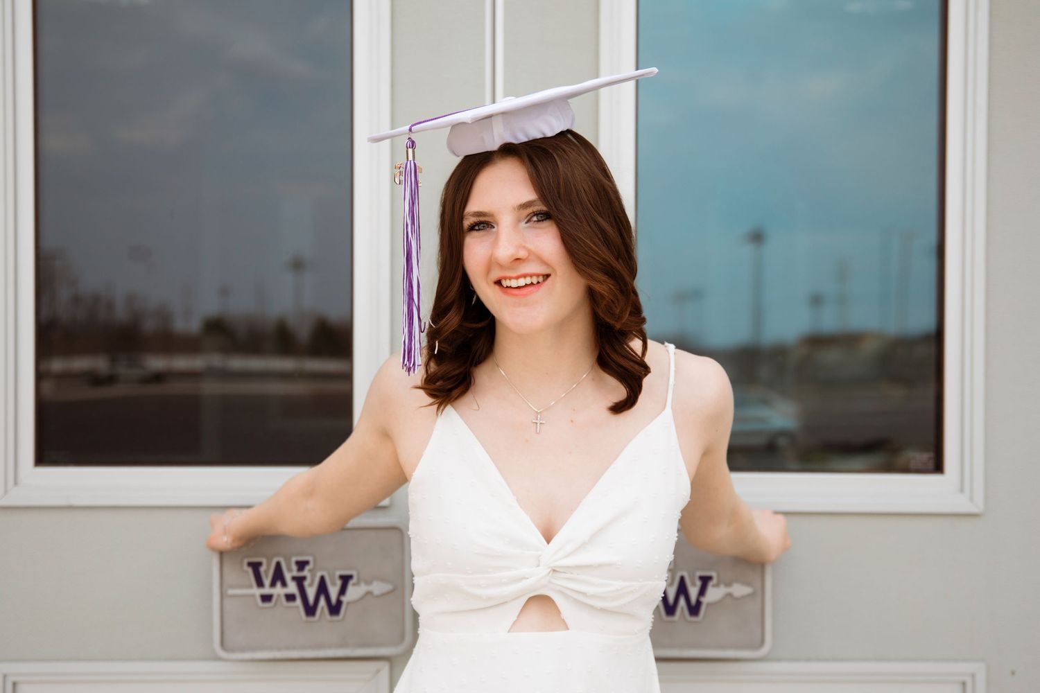 Young woman in white dress and lavender graduation cap smiling outdoors near a building with W logos.