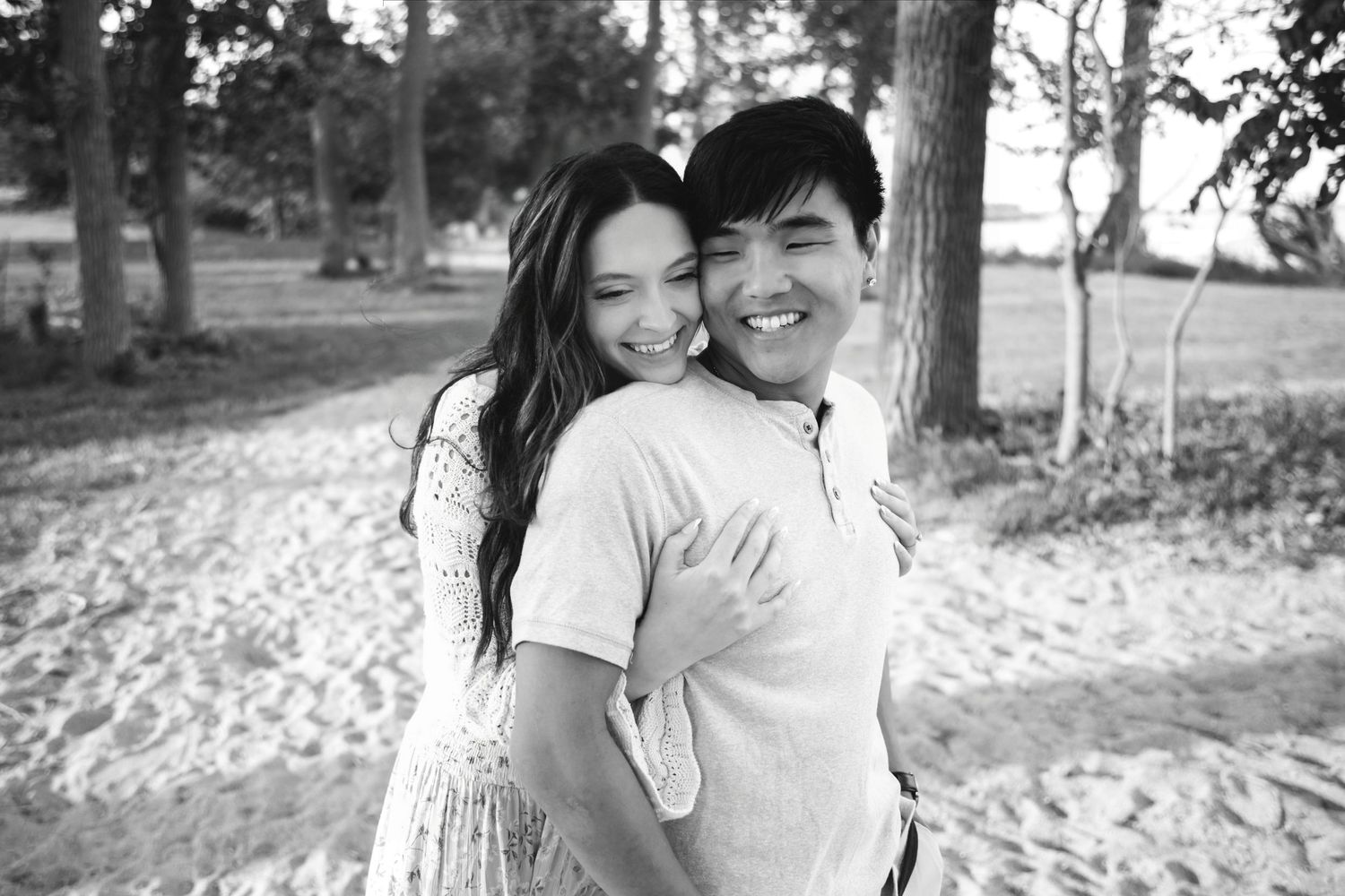 A joyful couple embraces and smiles outdoors on a sandy path surrounded by trees in a black and white photo.