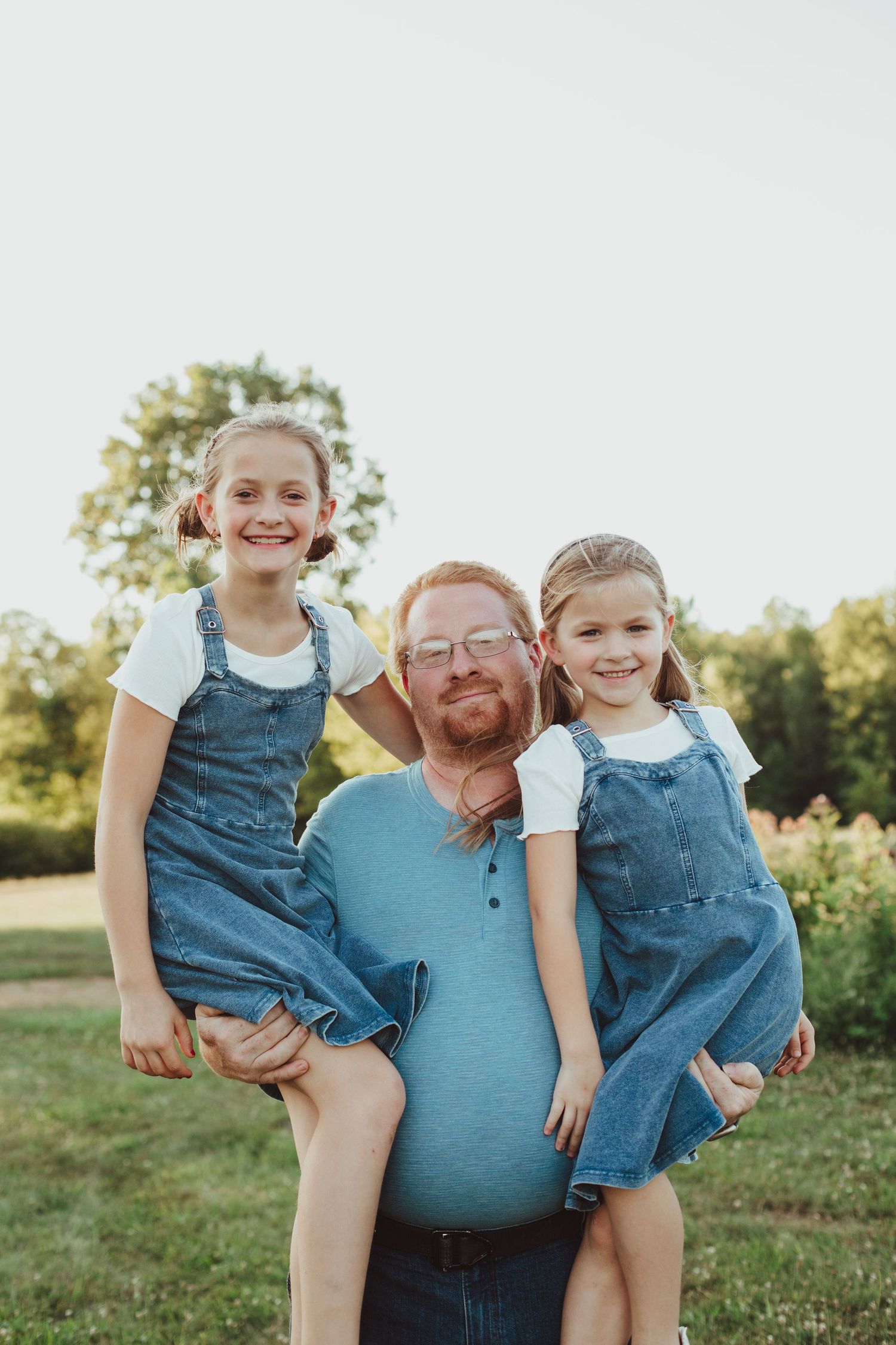 A bearded father with glasses holds two young girls in denim overalls in a sunny outdoor field.