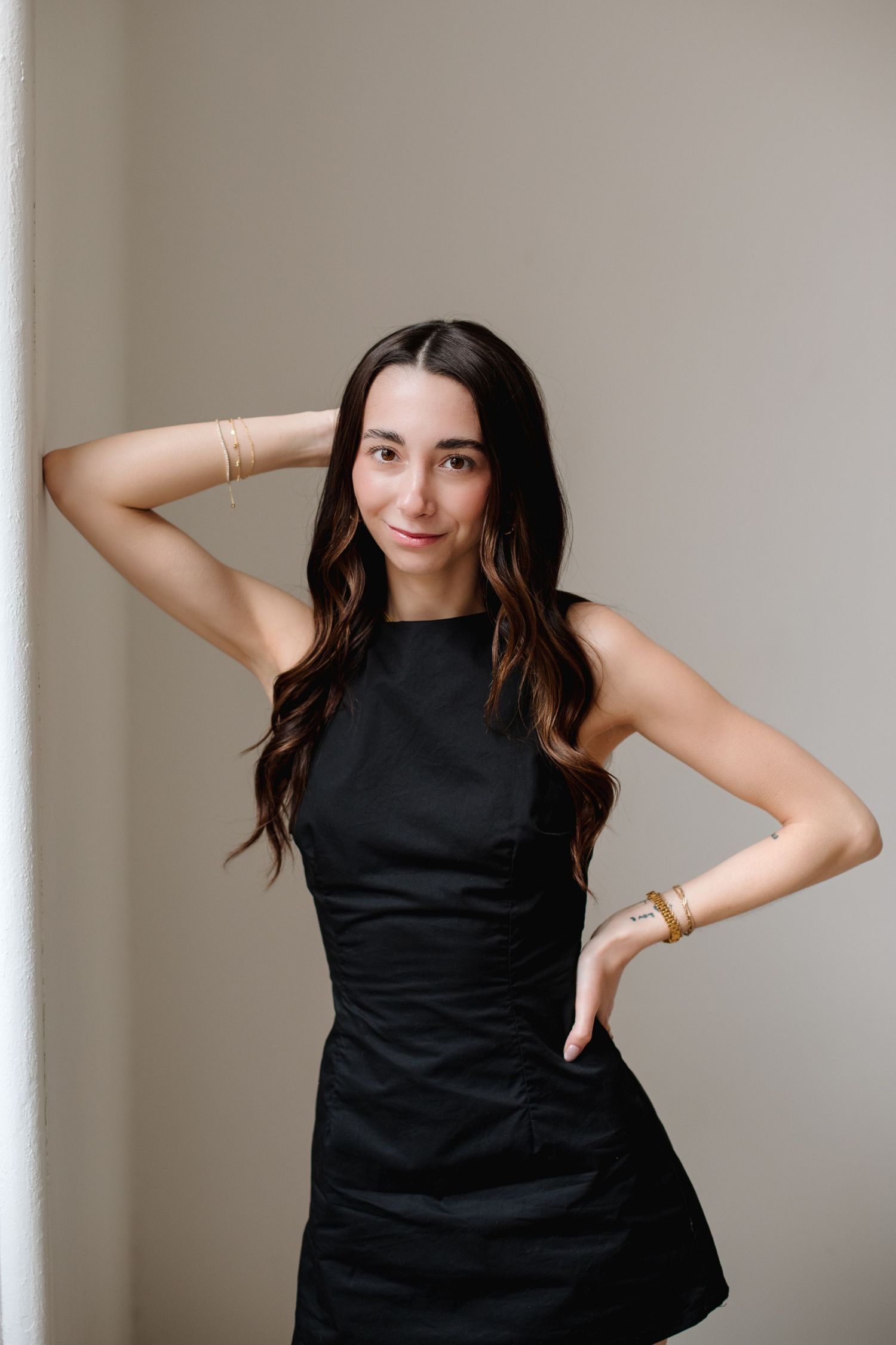 Young woman in black sleeveless dress posing with hand behind head near wall.