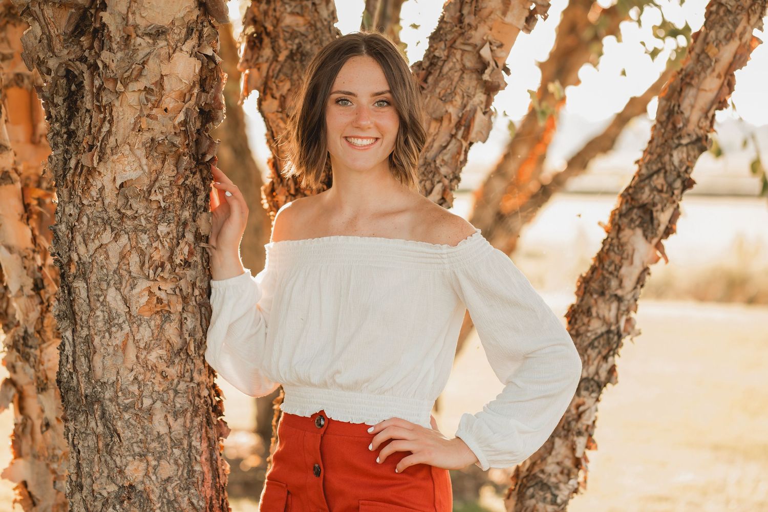 Happy young woman in white off-shoulder blouse and red skirt posing by rustic trees.