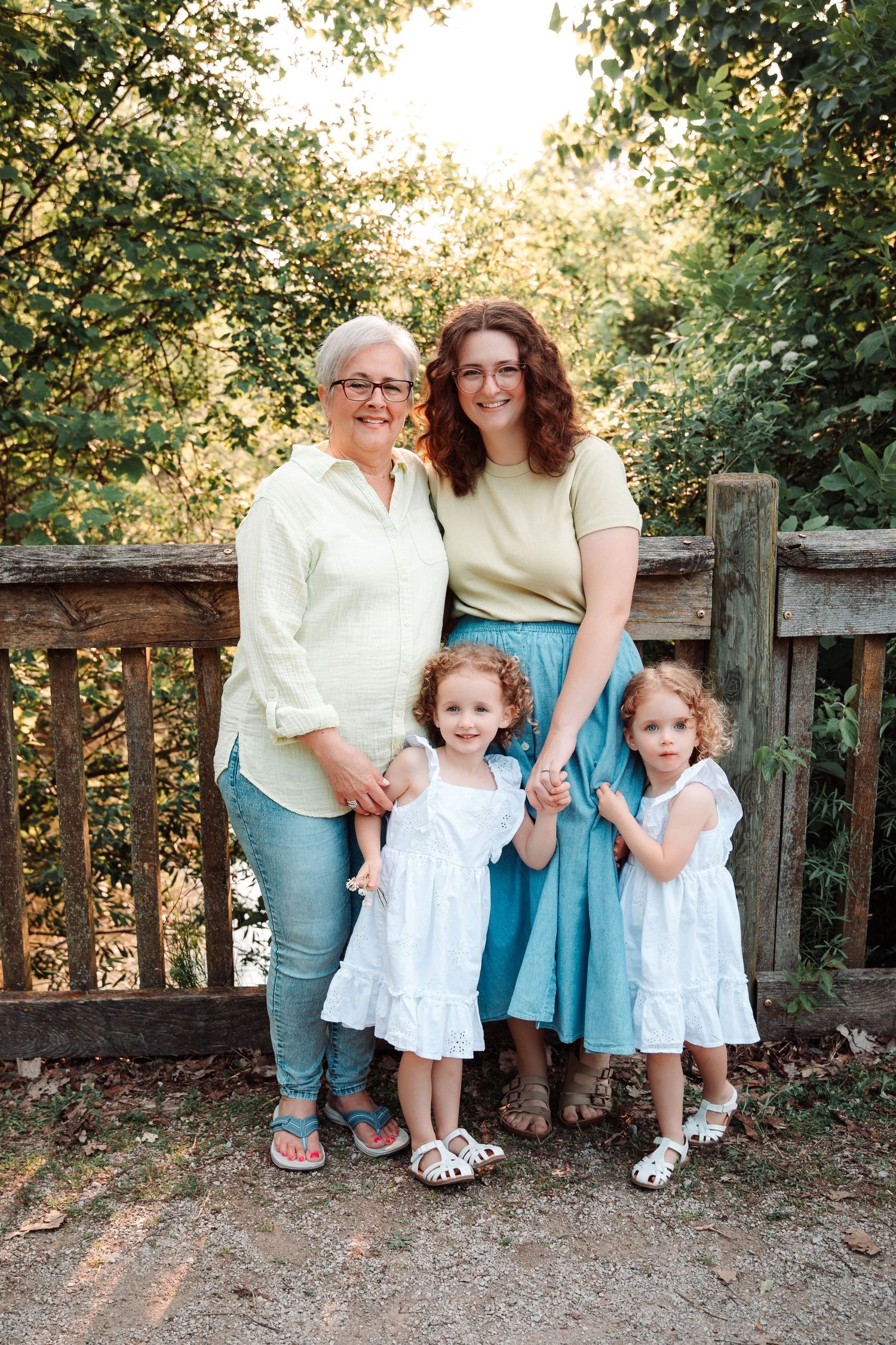 Two women and two young girls in white dresses pose together near a wooden fence outdoors.