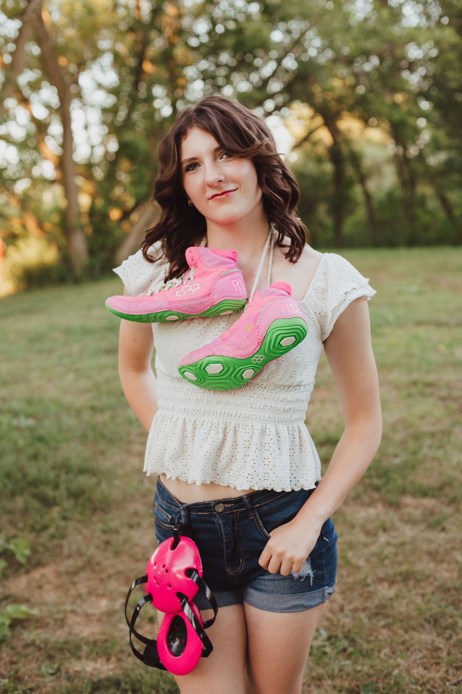 Young woman in white top and denim shorts holding pink and green sneakers outdoors in a park setting.
