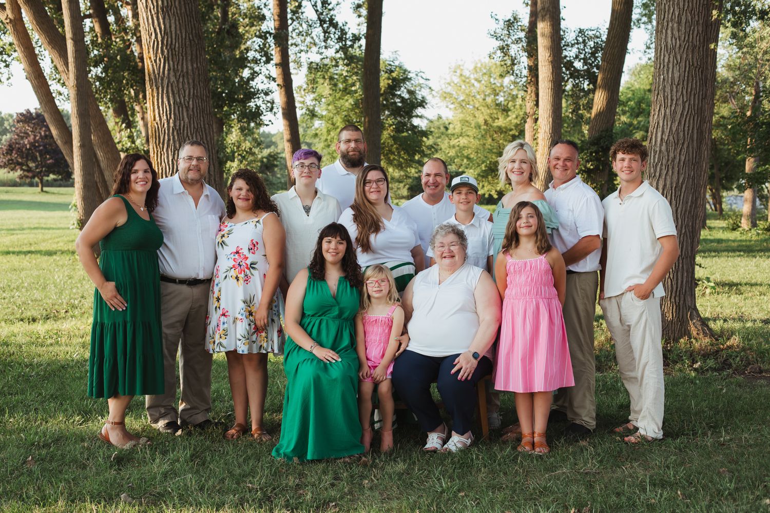 Large family group portrait outdoors in park with trees, wearing coordinated green, white, and pink outfits.