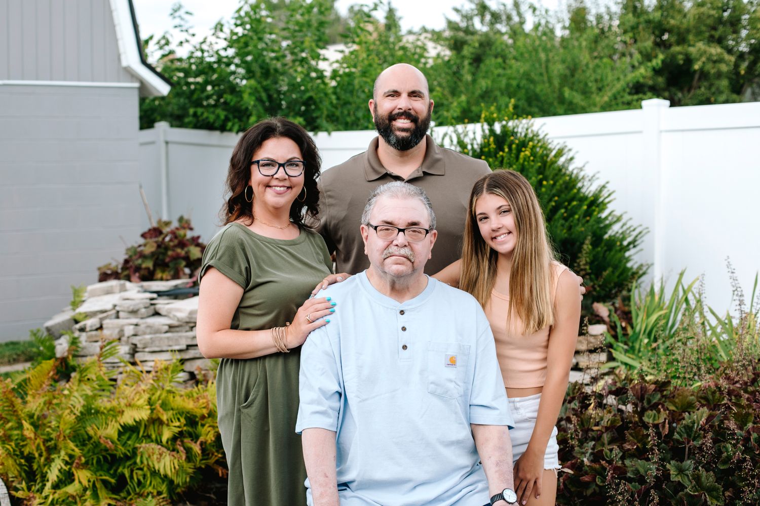 Family of four standing with elderly man in wheelchair in garden setting.