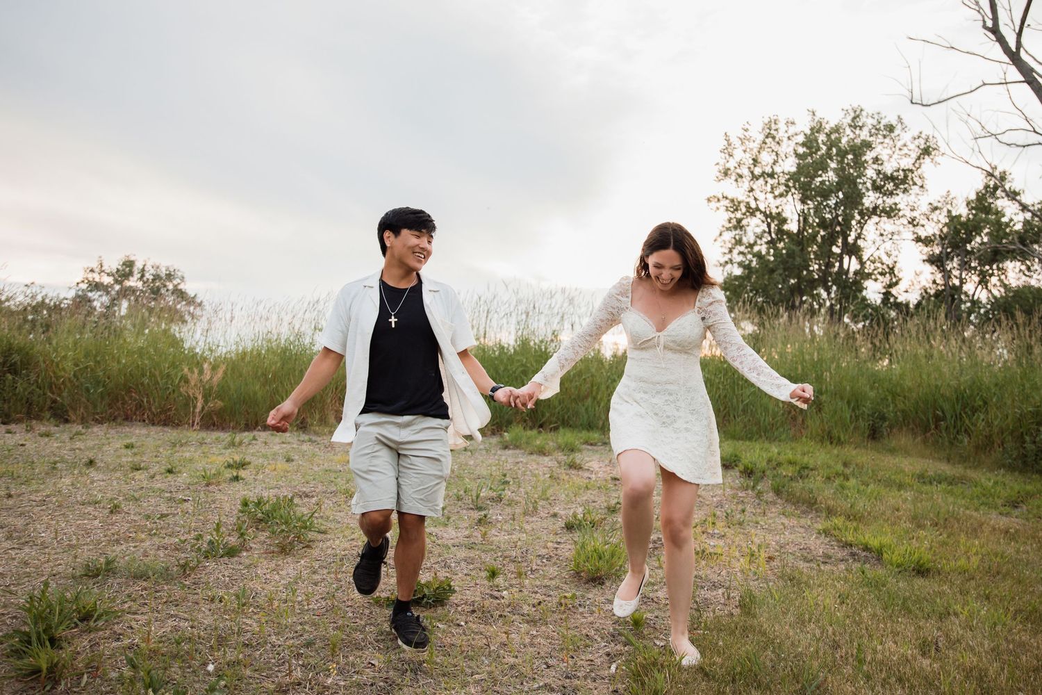 A happy couple runs hand in hand through an open field with tall grass under a cloudy sky.
