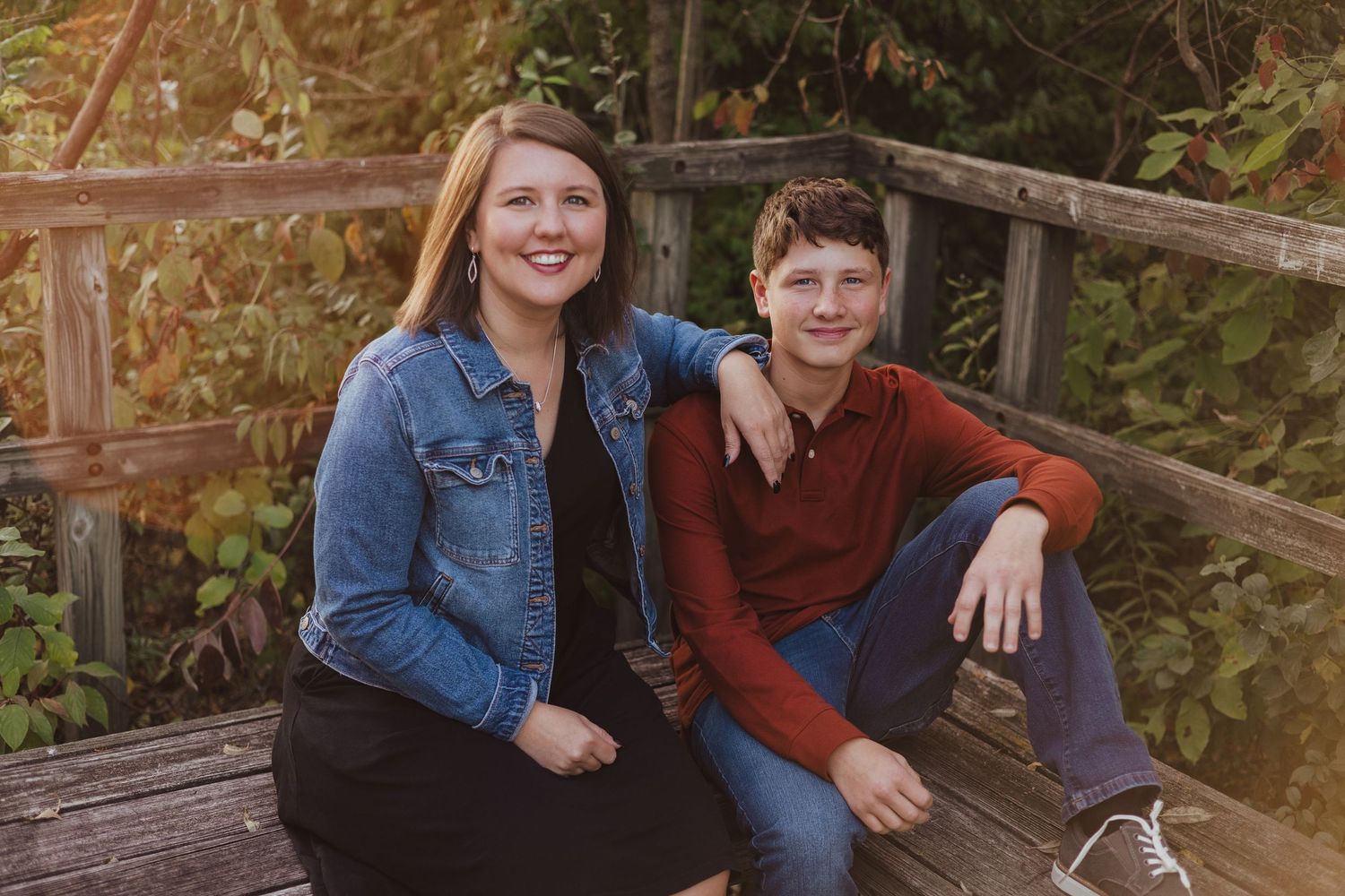 A smiling woman and teenage boy sitting together on a wooden bridge outdoors.