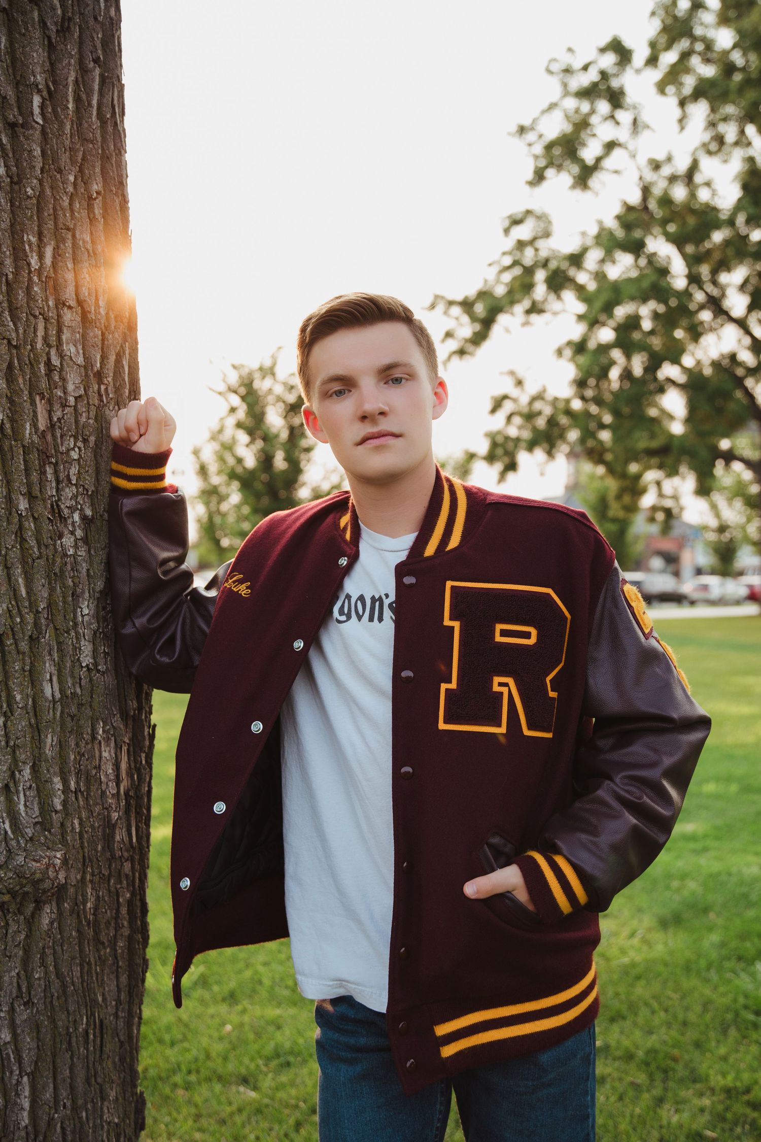 Young man in maroon varsity jacket with R letter leaning against tree at sunset in park.