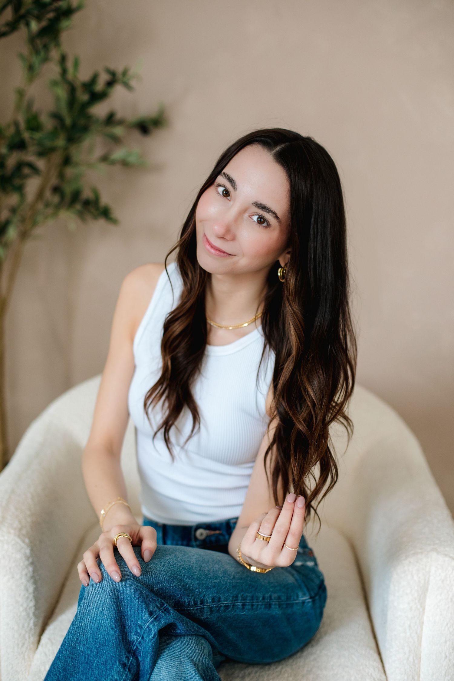 Young woman with long dark hair sitting in white chair wearing white tank top, gold jewelry, and blue jeans.