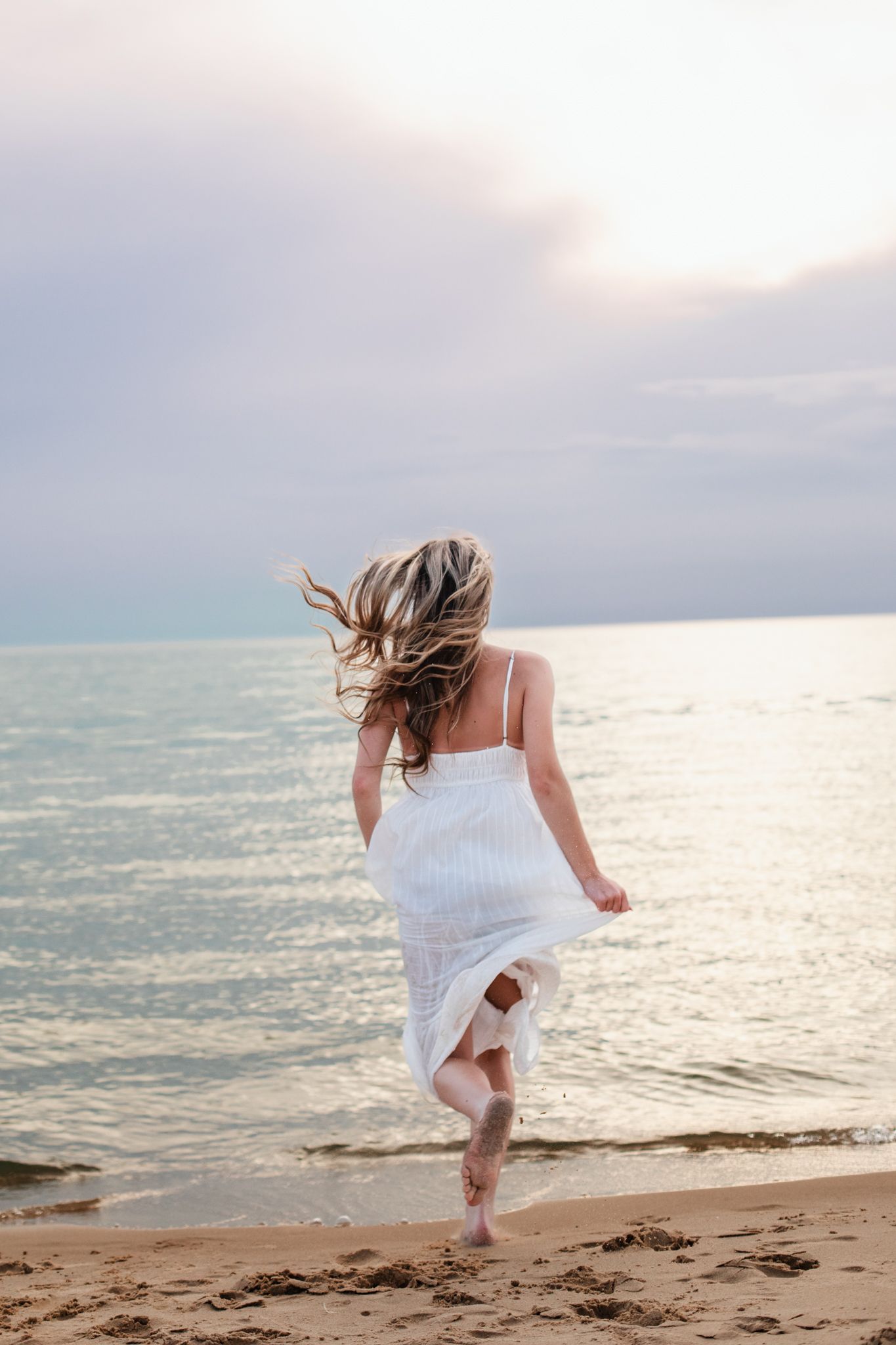 Young woman in white dress running barefoot on sandy beach toward calm ocean at sunset.
