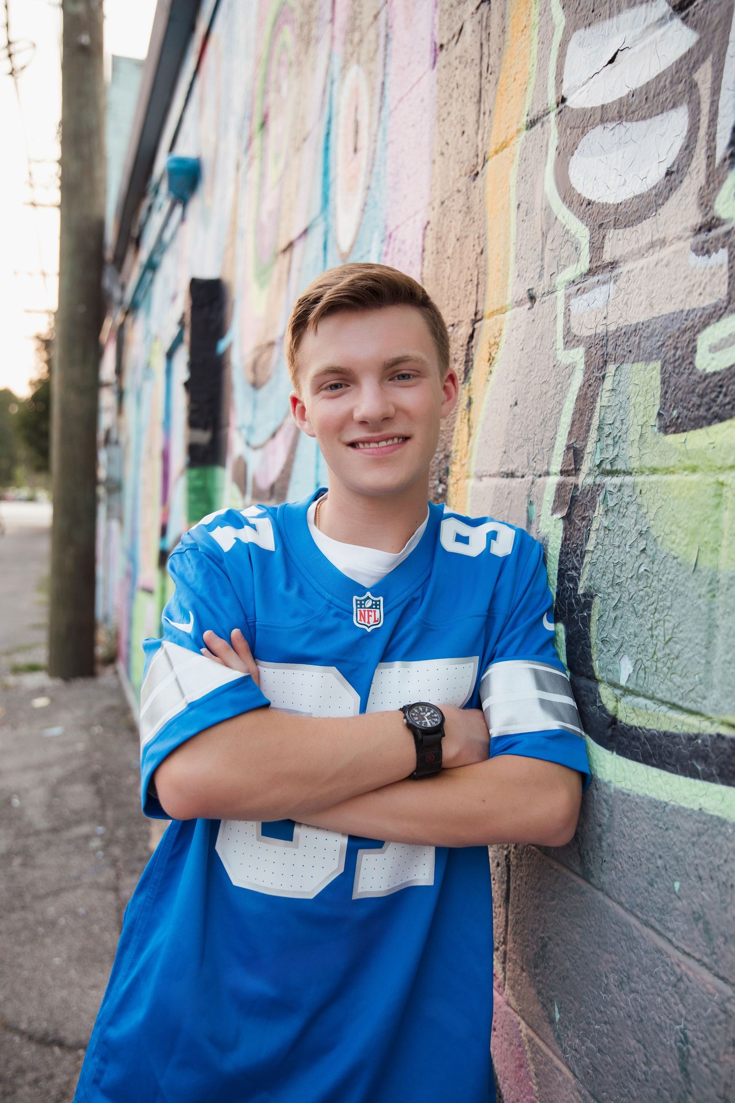 Teen in blue Lions NFL jersey smiling with arms crossed against graffiti covered brick wall.