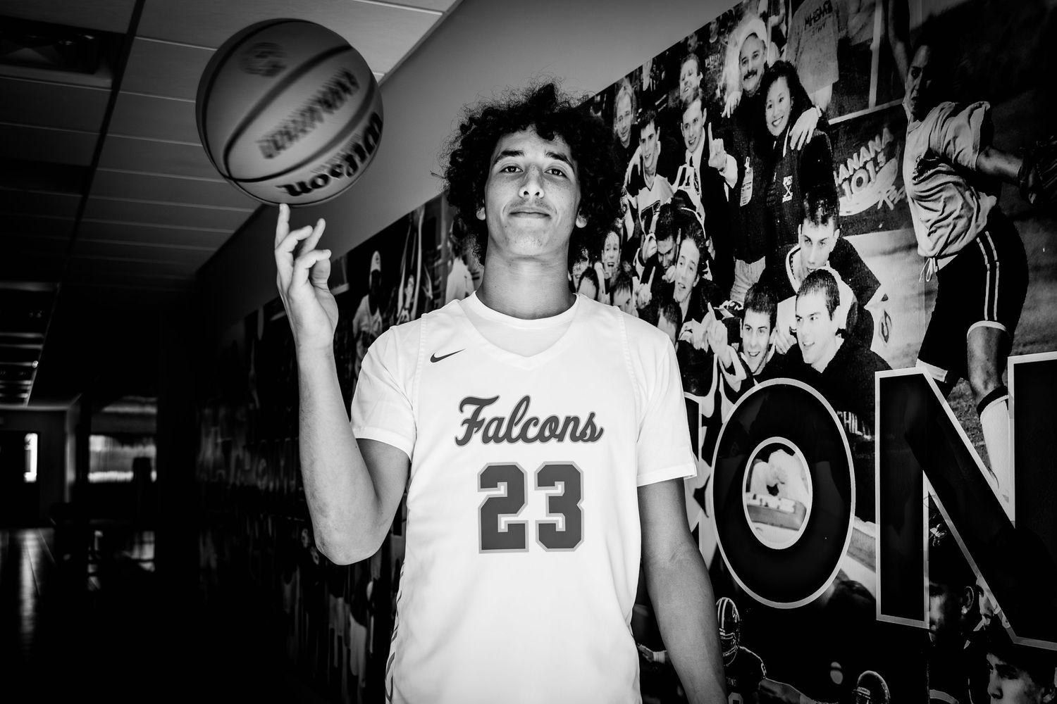 Black and white photo of a teen in a Falcons 23 jersey spinning a basketball on his finger in a school hallway.