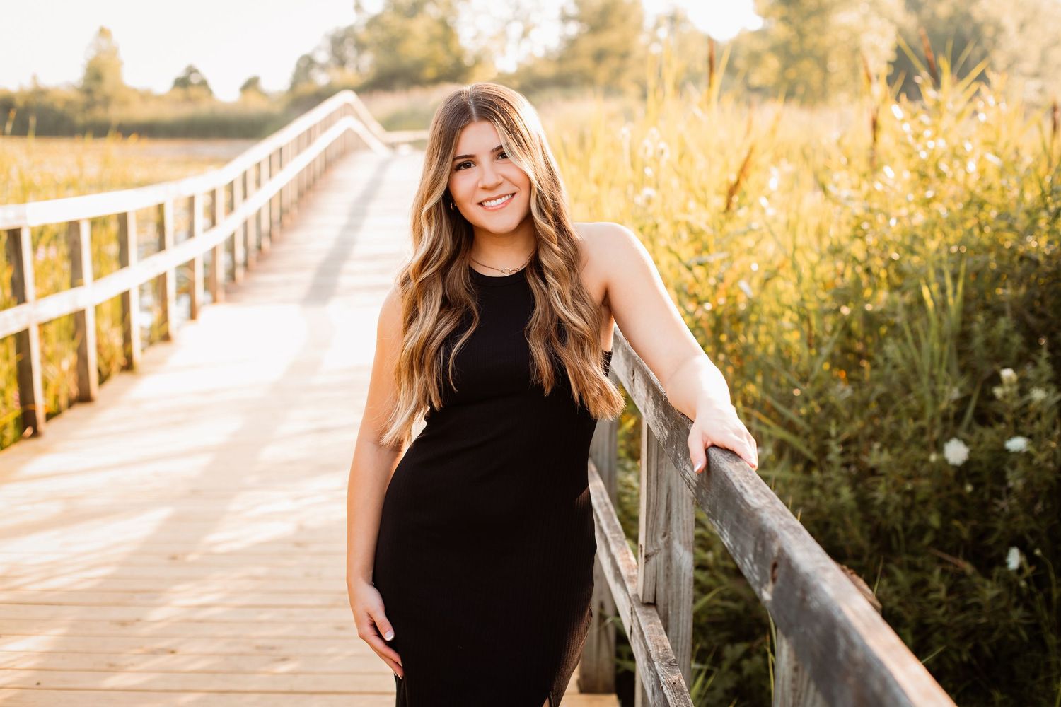 Young woman in black dress posing on wooden boardwalk surrounded by golden meadow grasses in warm sunlight.