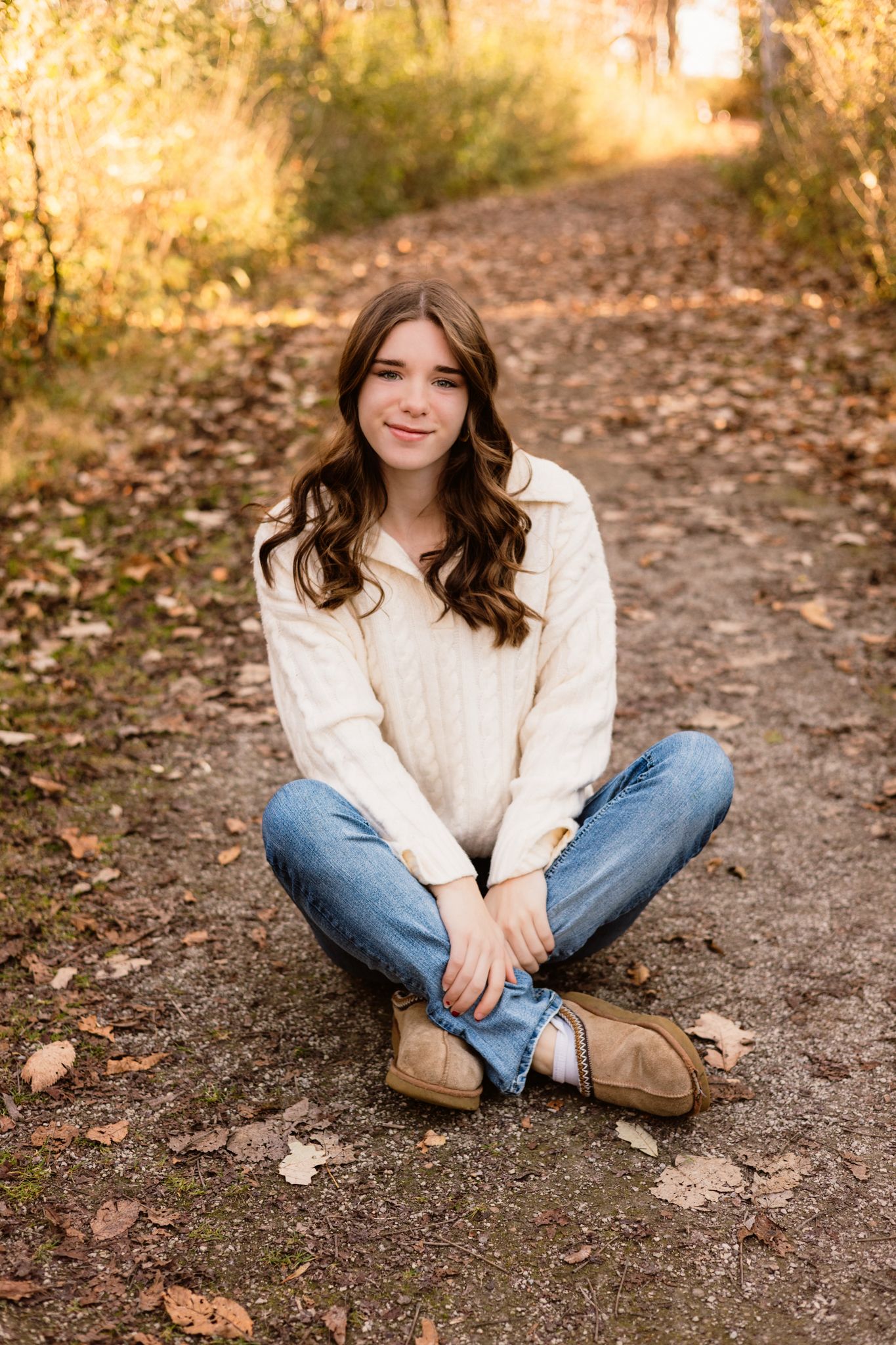 Teen girl in white cable knit sweater and jeans sits cross-legged on autumn forest path.
