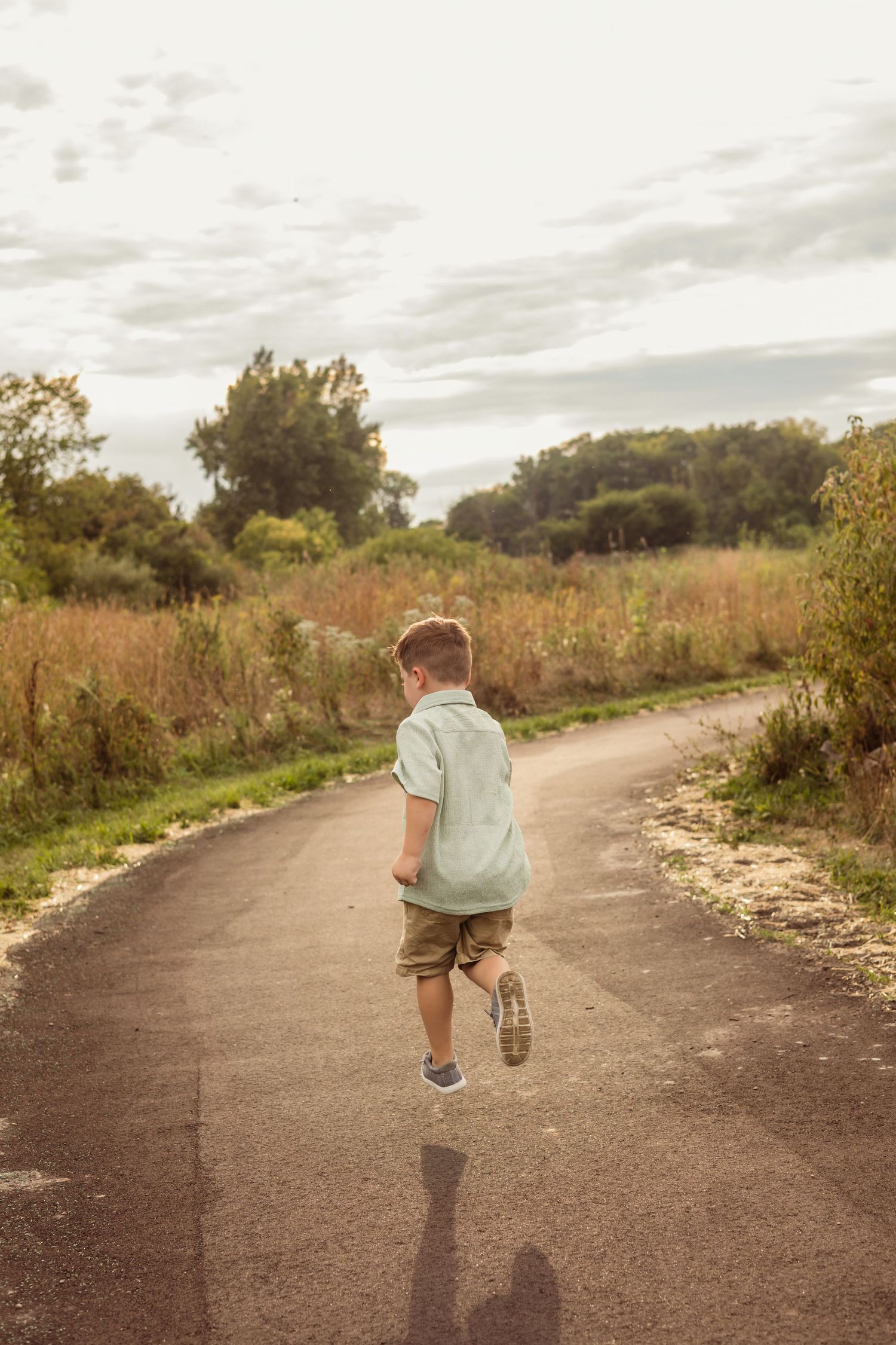 Young boy running away on a paved path surrounded by green fields and trees under a cloudy sky.