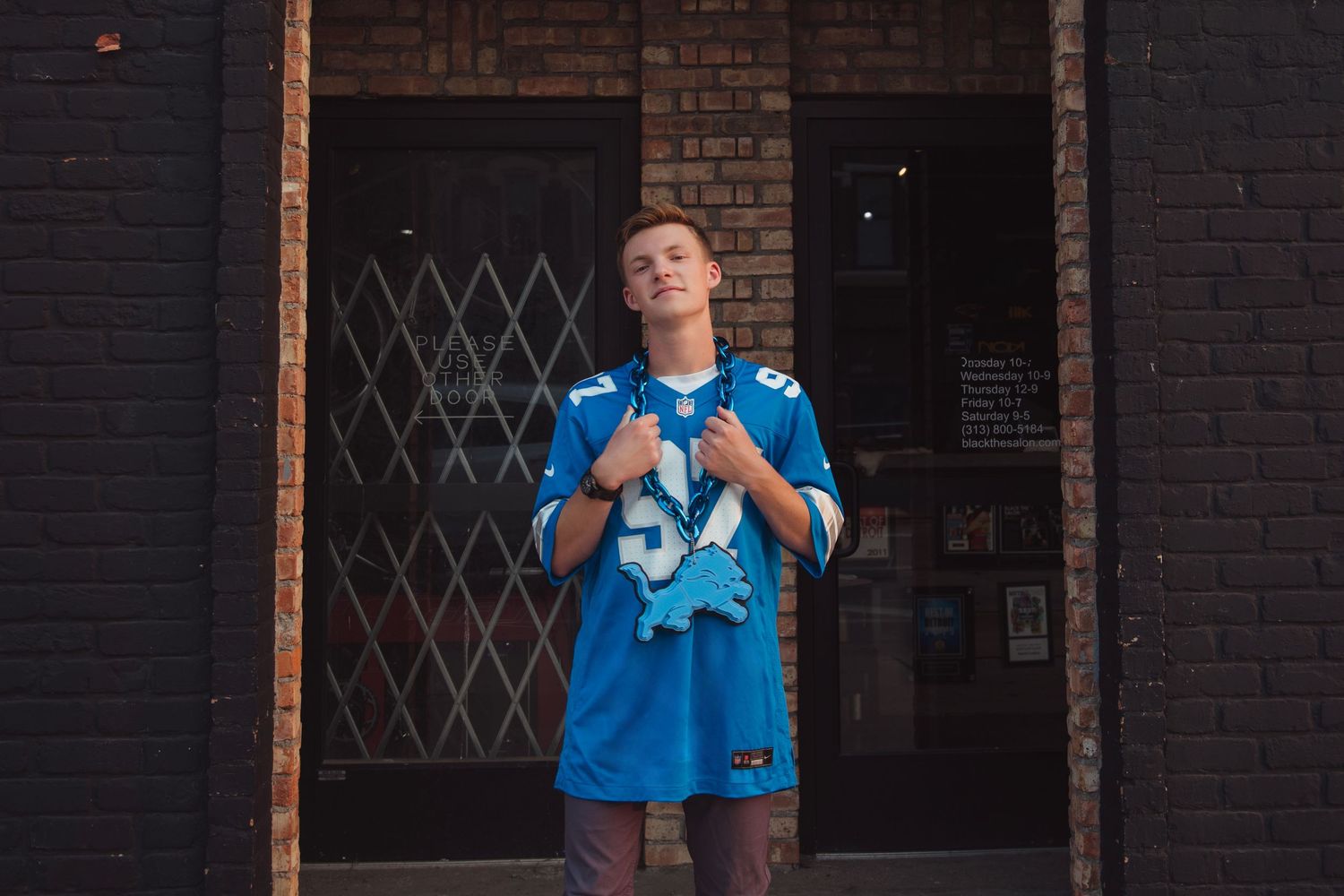 Teen boy in blue Detroit Lions NFL jersey number 97 posing by brick building entrance.