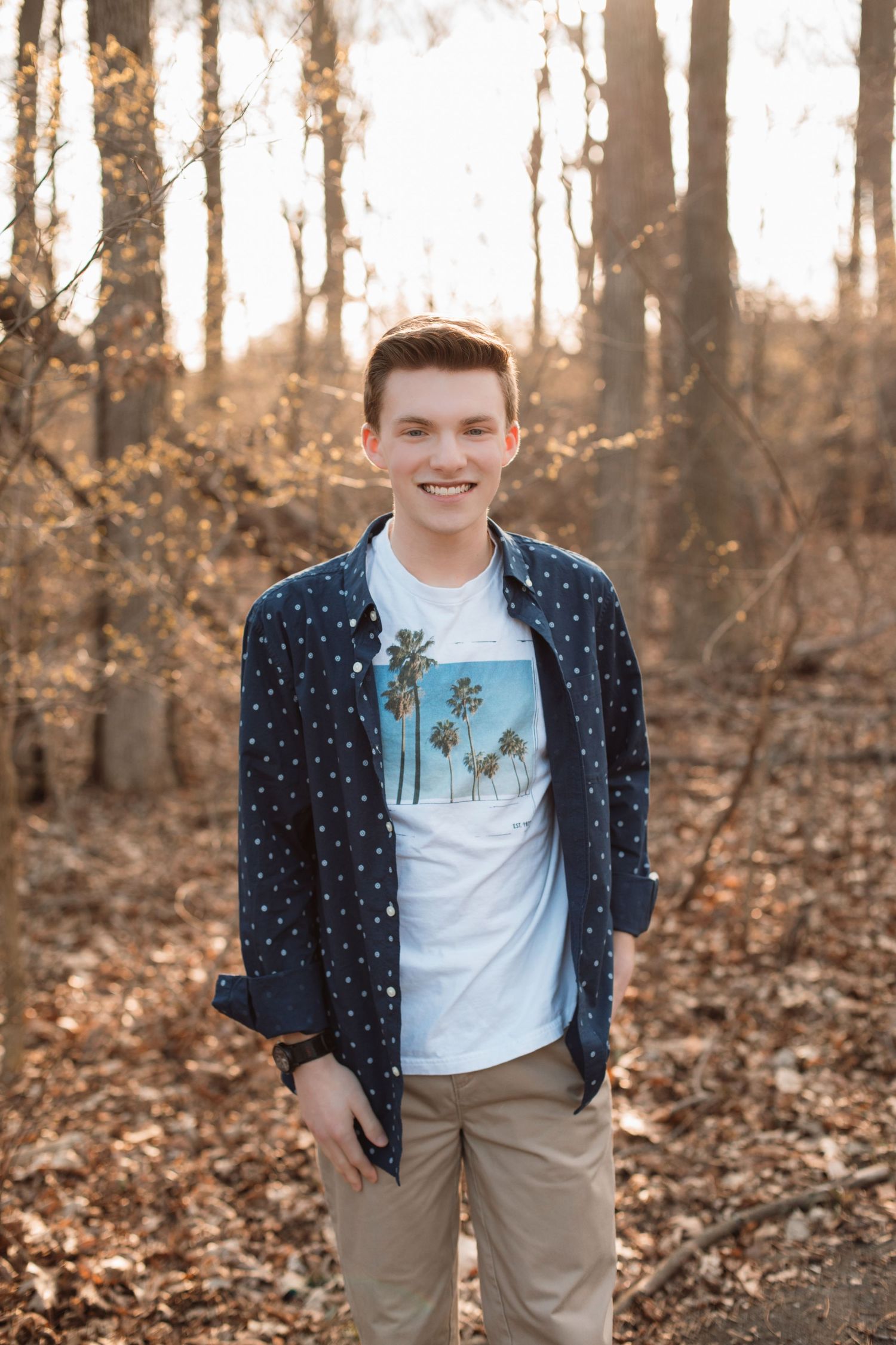 Happy teen boy in navy polka dot shirt and palm tree graphic tee stands in sunlit autumn forest.