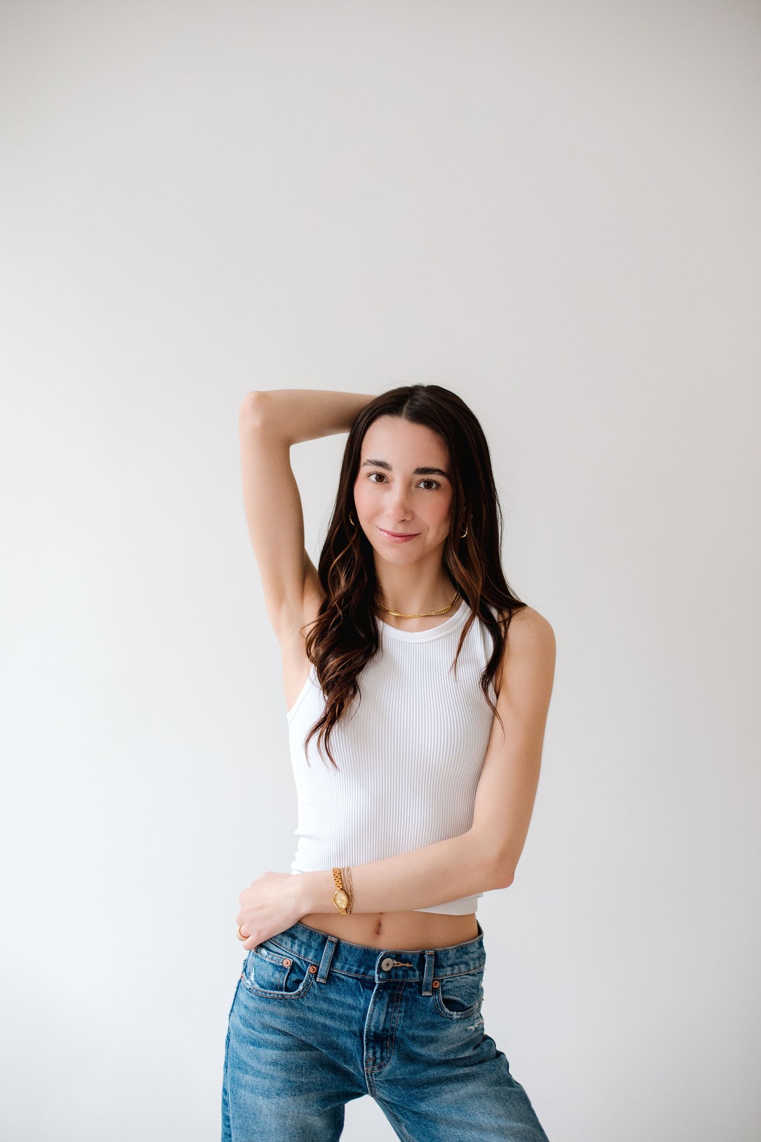Young brunette woman in white crop top and jeans posing with one arm raised against white background.