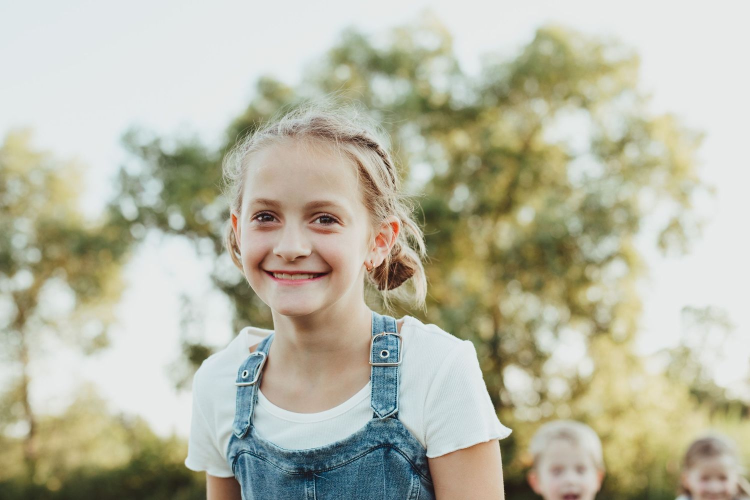 A smiling young girl with braids in denim overalls poses outdoors in soft natural golden light.