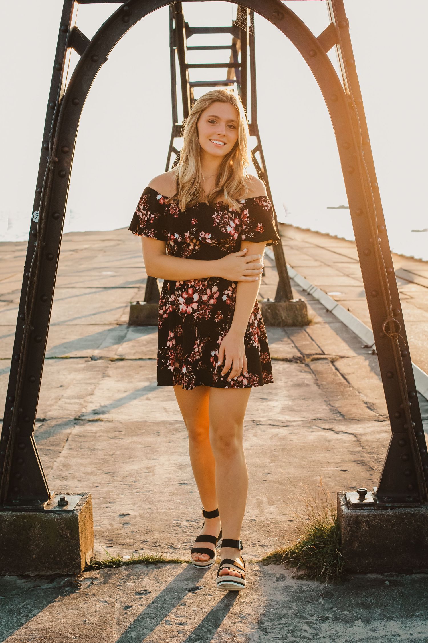 Young blonde woman in floral off-shoulder dress smiling under metal arch structure at sunset.