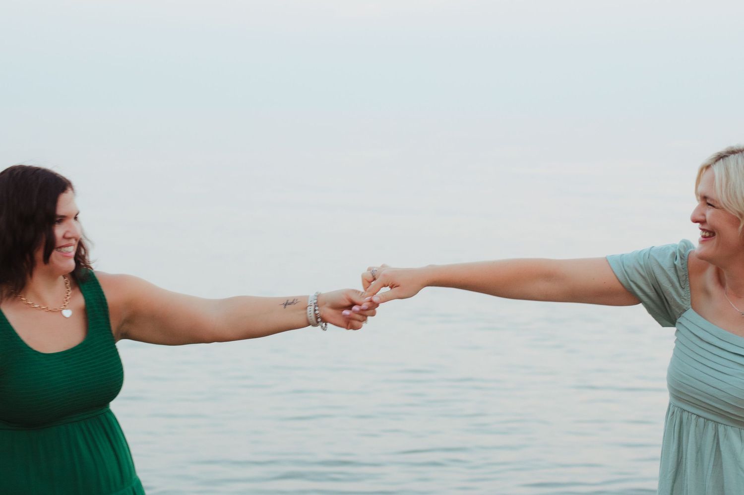Two women laughing and holding hands at arms length with ocean in background.