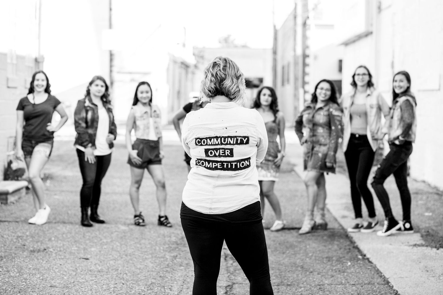 Woman wearing Community Over Competition jacket facing group of women in alley, black and white.