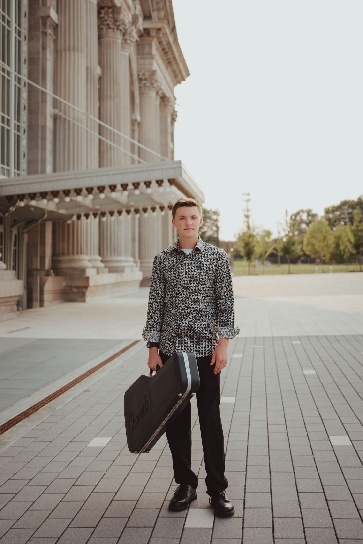 Young man in patterned shirt holding instrument case outside classical building with columns.