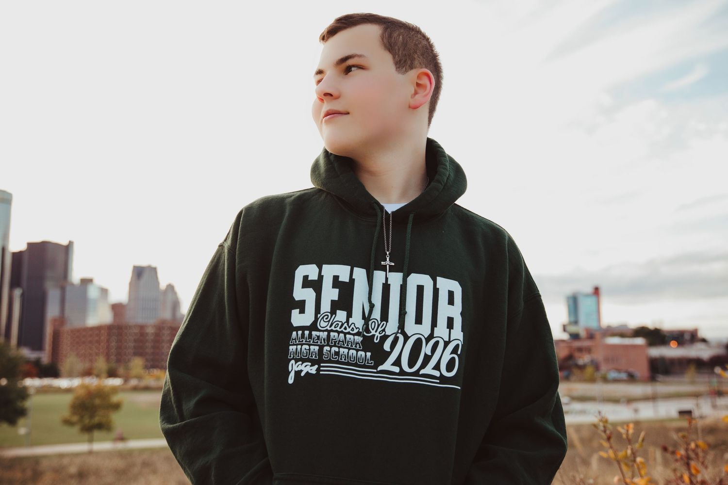 Teen wearing a black Senior 2026 hoodie sweatshirt posing outdoors with a city skyline in the background.