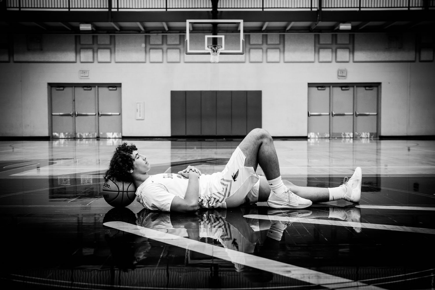 Black and white photo of a basketball player lying on a gym floor with a ball, hoop visible in background.