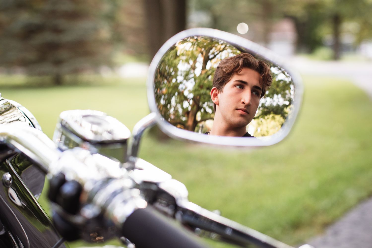 Young man's reflection visible in a chrome motorcycle side mirror with green trees in the background.