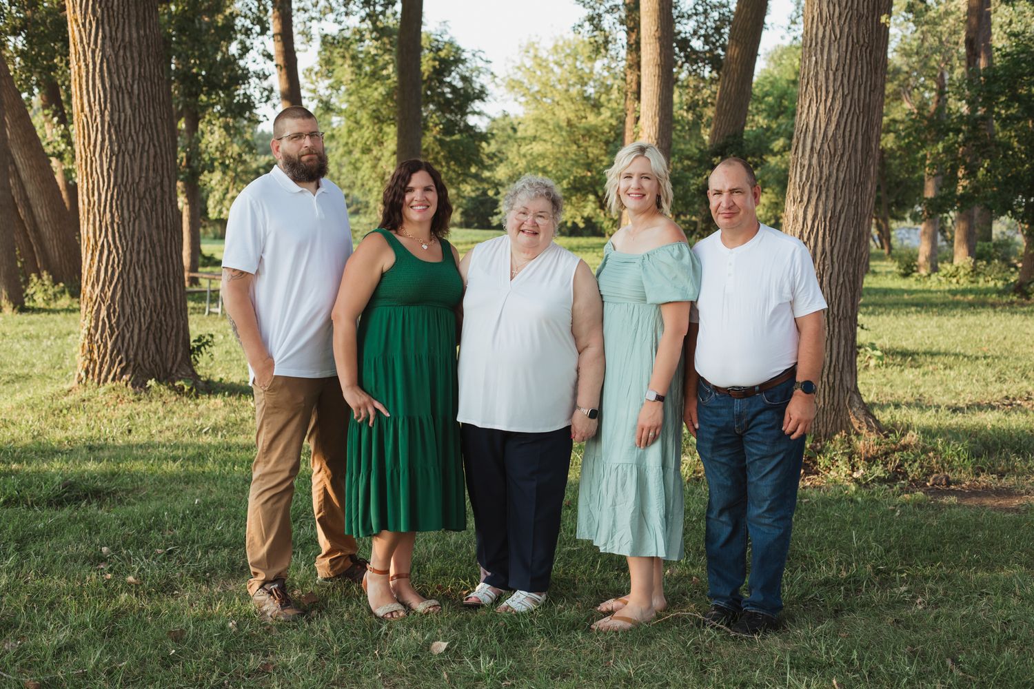 Family of five smiling outdoors in a park, wearing green and white outfits, surrounded by tall trees.