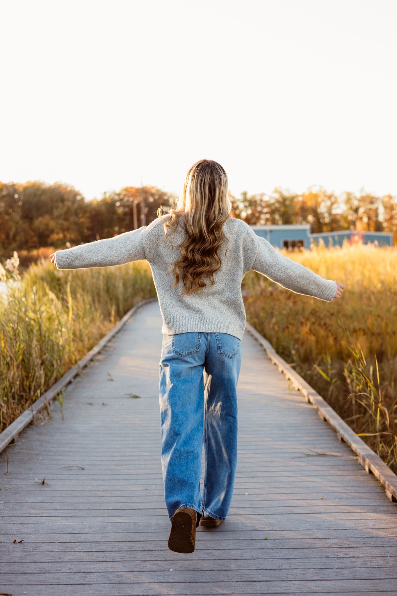 Woman in cream sweater and wide-leg jeans walks along wooden boardwalk with arms outstretched in golden autumn light.