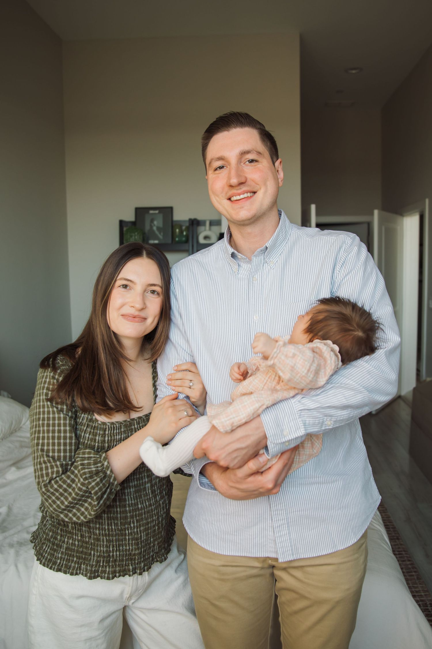 Smiling parents holding newborn baby together in bright bedroom at home.