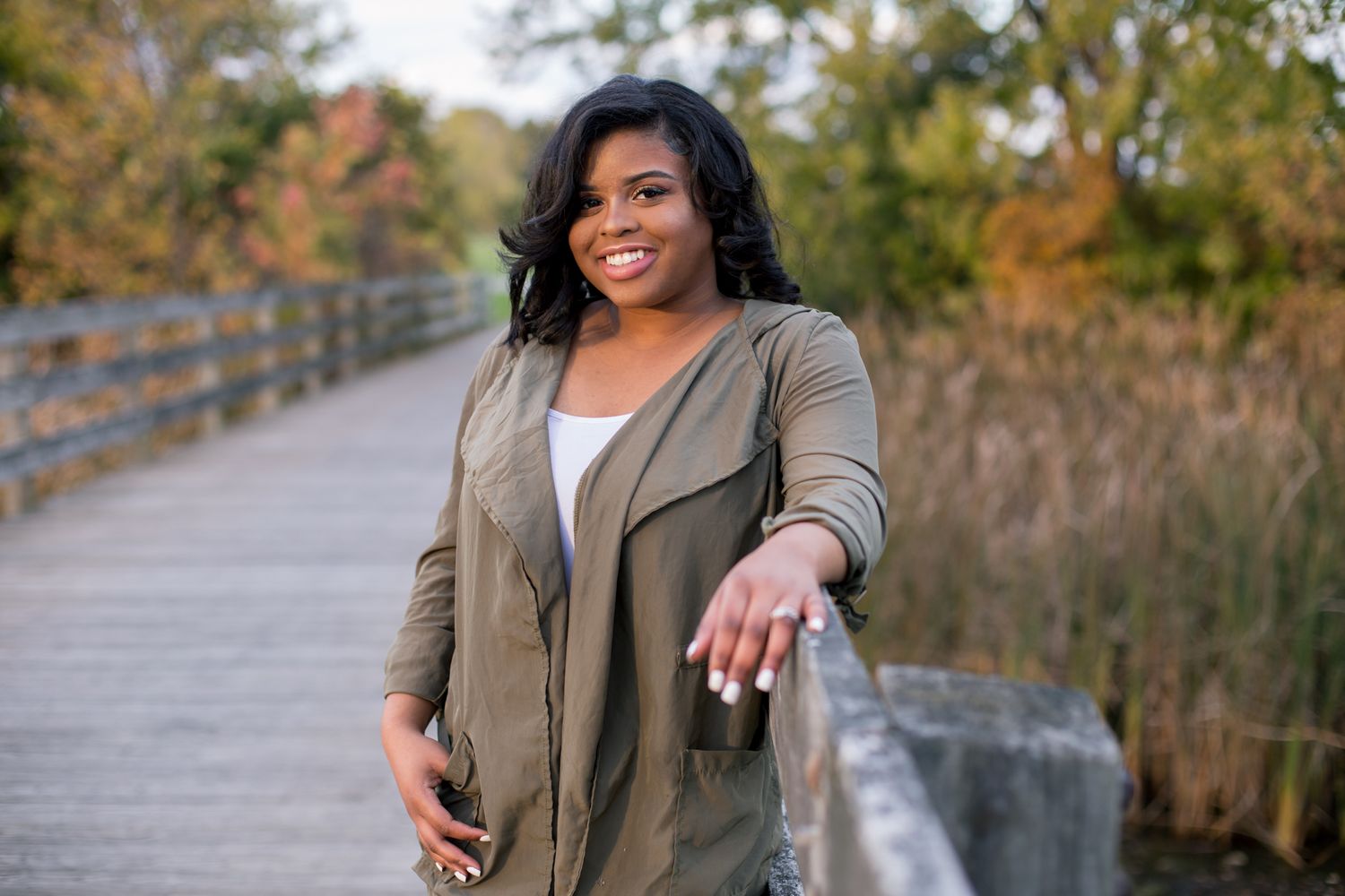 Smiling woman in olive green jacket posing on wooden boardwalk surrounded by autumn foliage.