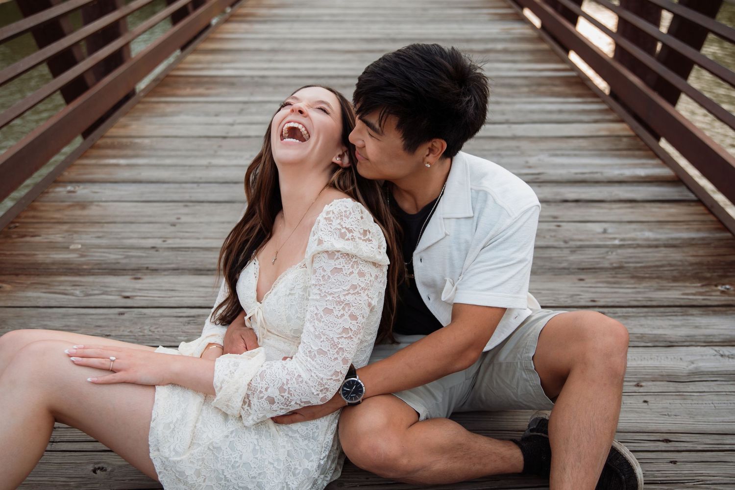 Couple laughing joyfully while sitting on a wooden bridge, sharing a playful and loving moment.