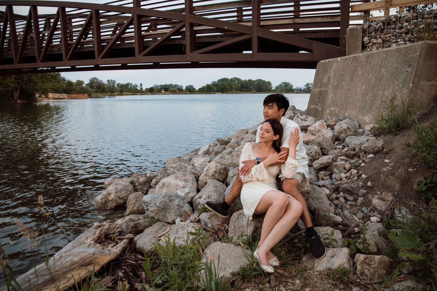 Couple sitting together on rocky shoreline under a bridge near calm water, sharing a quiet romantic moment.