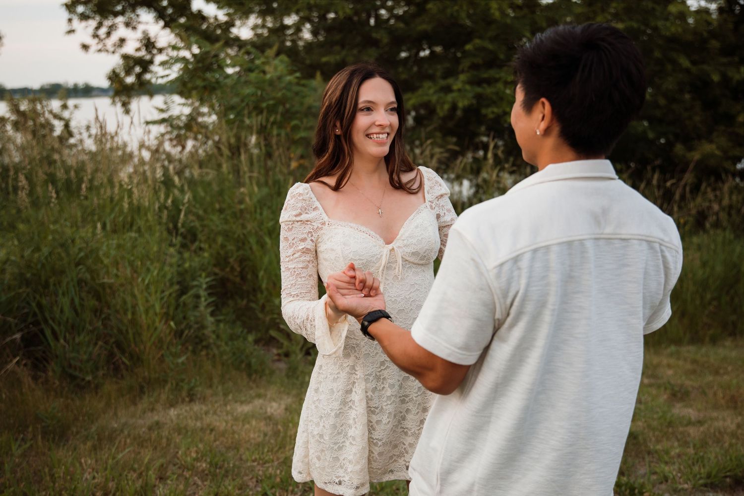 Smiling woman in white lace dress holds hands with man near tall grass and lake in soft evening light.